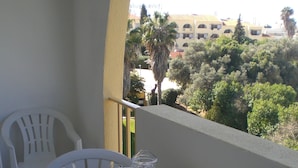 Un balcón con una pequeña mesa y dos sillas, una jarrón de vidrio con una bebida, y vistas de un edificio y árboles.