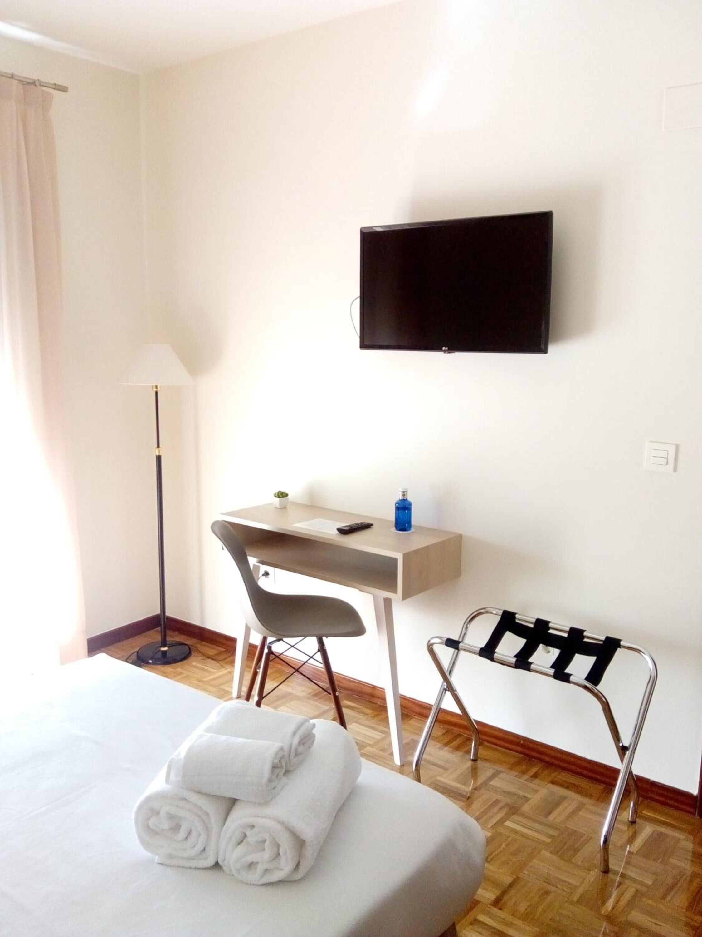 A hotel room with a bed, a desk, a chair, a TV mounted on the wall, and a towel rack.