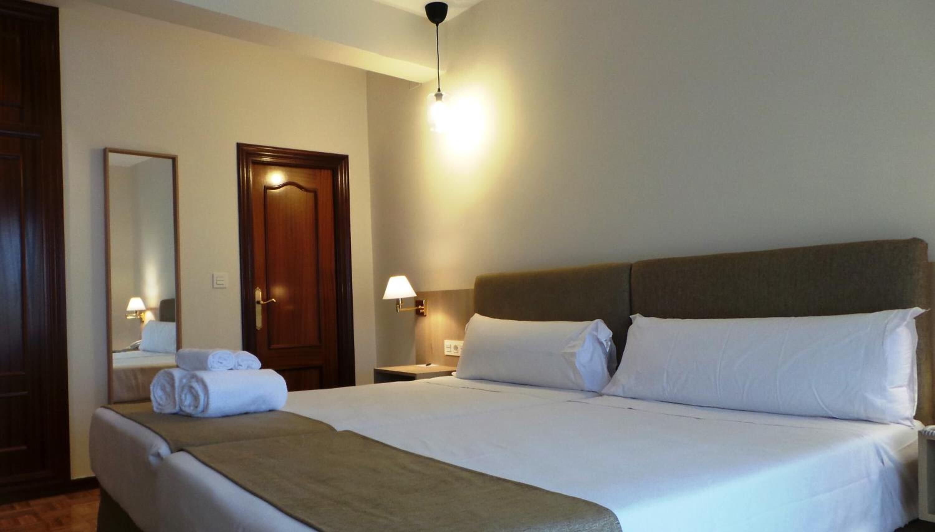 A hotel room with a large bed, a wooden door, a mirror, and a nightstand with a lamp.