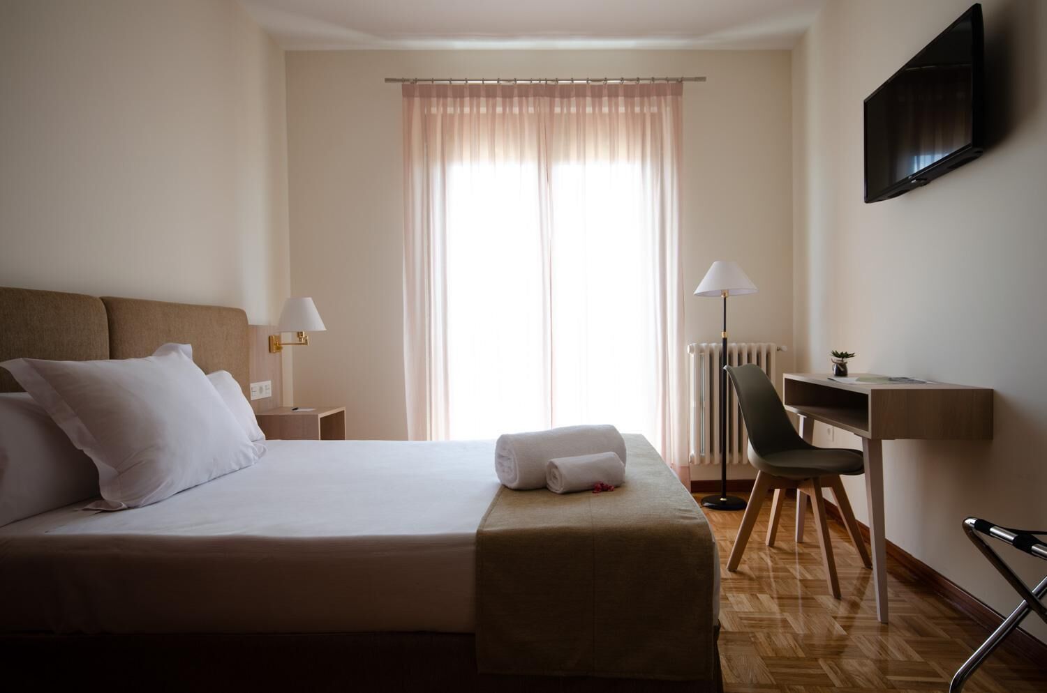Habitación de hotel con cama, escritorio, silla, televisor y ventana con cortinas.
