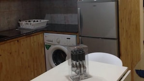 Een compacte keuken met houten kastjes, een wasmachine, een koelkast en een kleine tafel met een doorzichtige container erop.