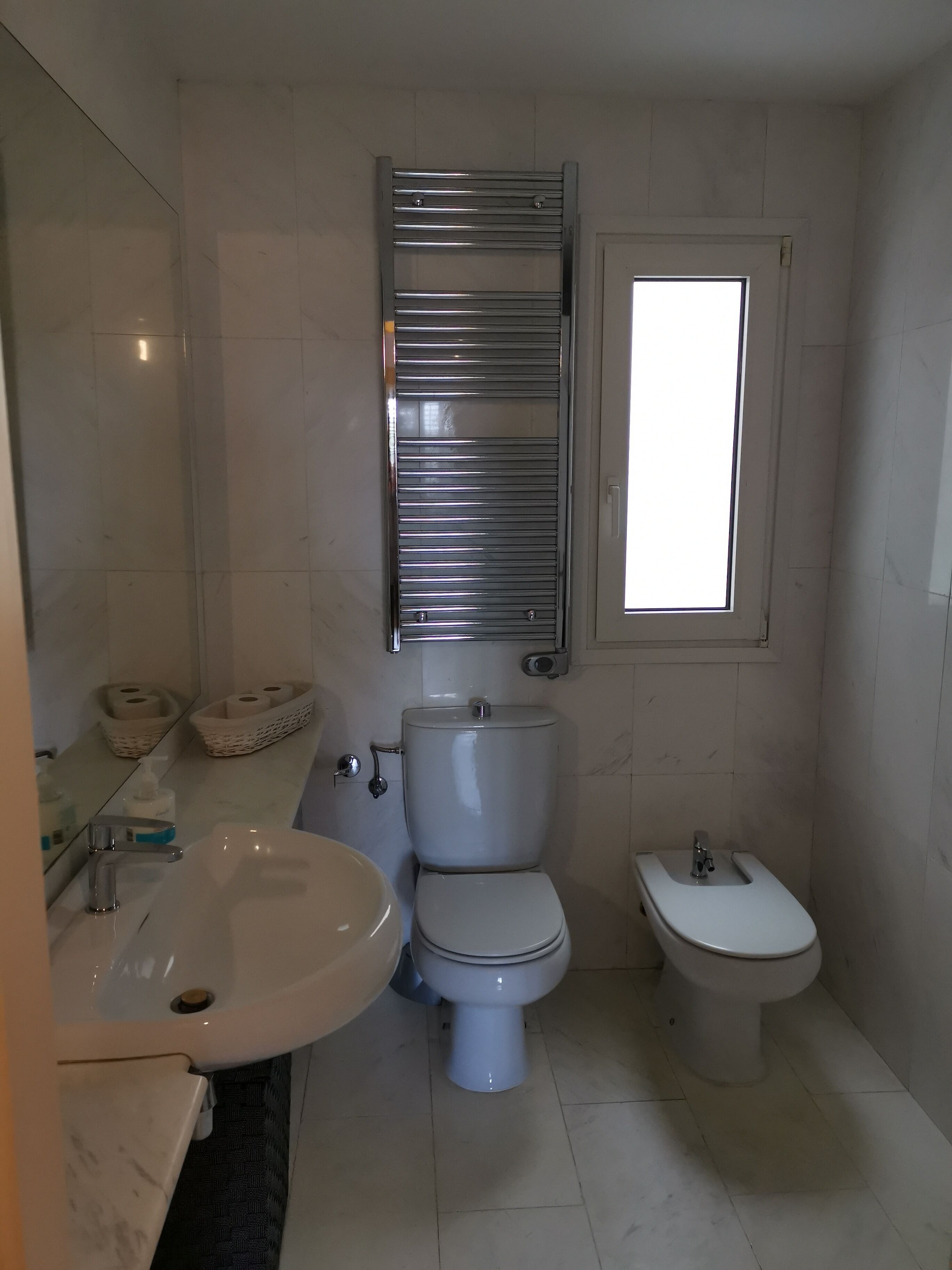 A bathroom with a sink, toilet, and bidet. There is a mirror above the sink and a window above the toilet.