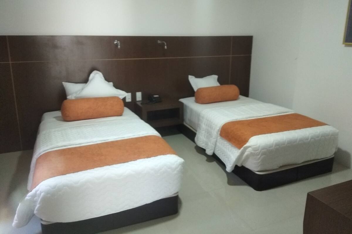 A hotel room with two beds, each with white bedding and orange pillows. There is a wooden headboard, a nightstand with a phone, and a small table.
