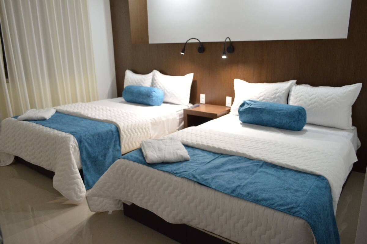 A hotel room with two beds, each with a blue blanket and white pillows. There are bedside tables with lamps and a large window with blinds.