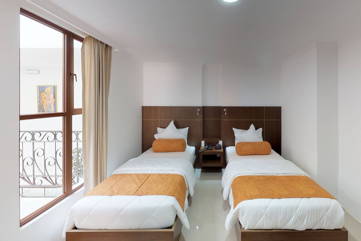 A hotel room with two beds, a nightstand, a window with curtains, and a view of the street.