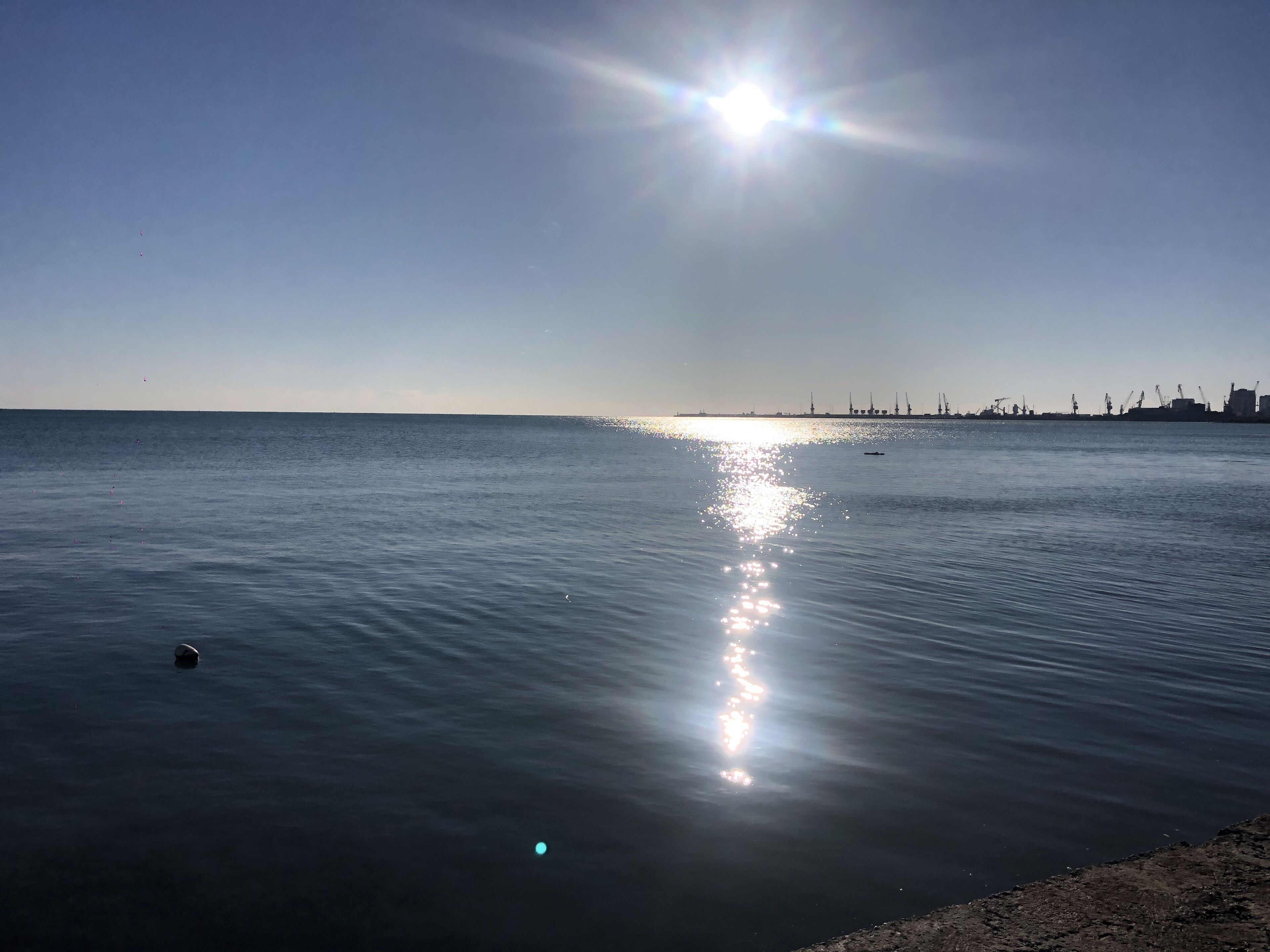 Ruhige See mit der Sonne, die sich im Wasser spiegelt, eine ferne Küste mit Gebäuden und einem klaren Himmel.