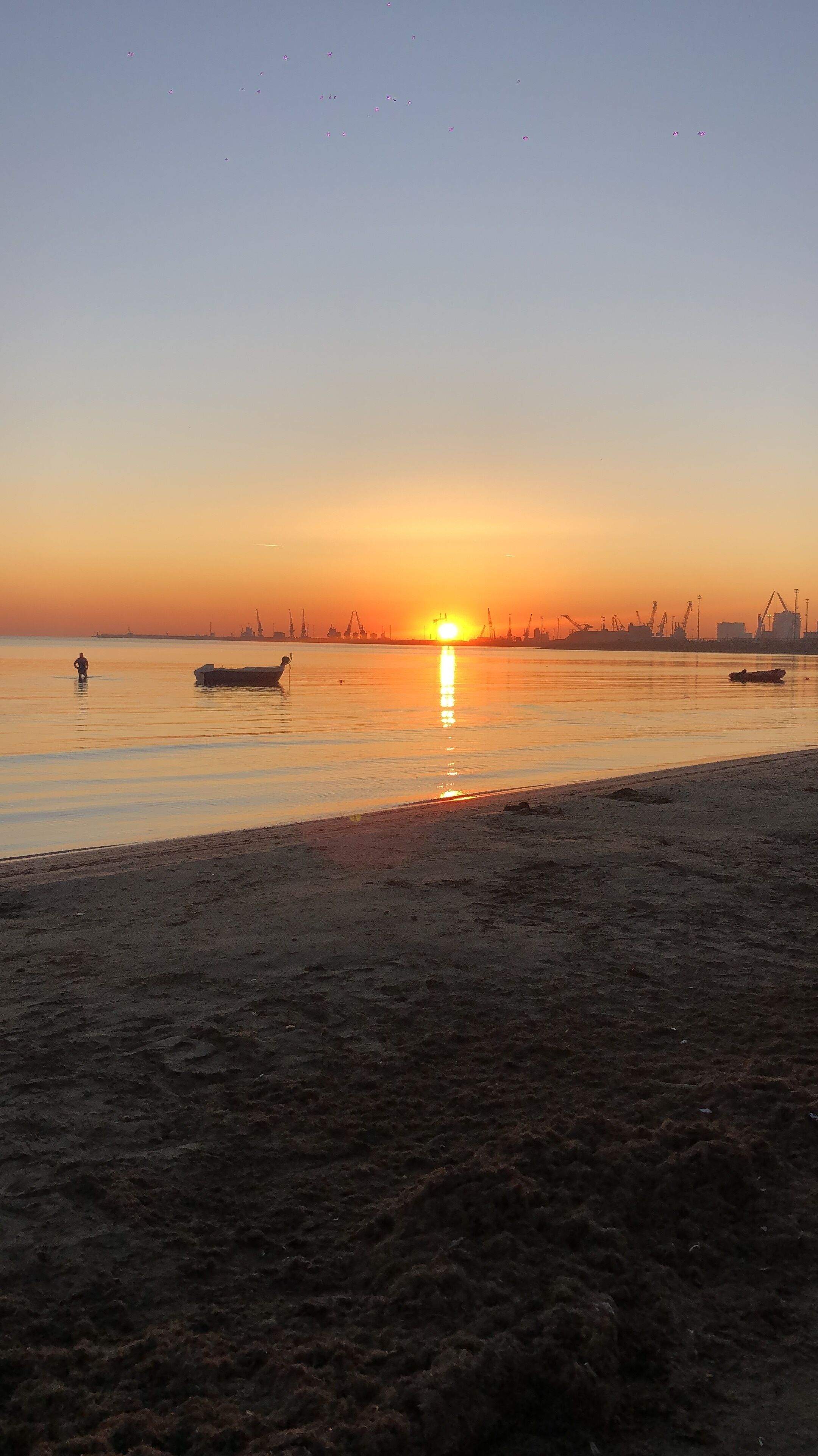 A sunset over a calm body of water with boats and a distant industrial skyline.