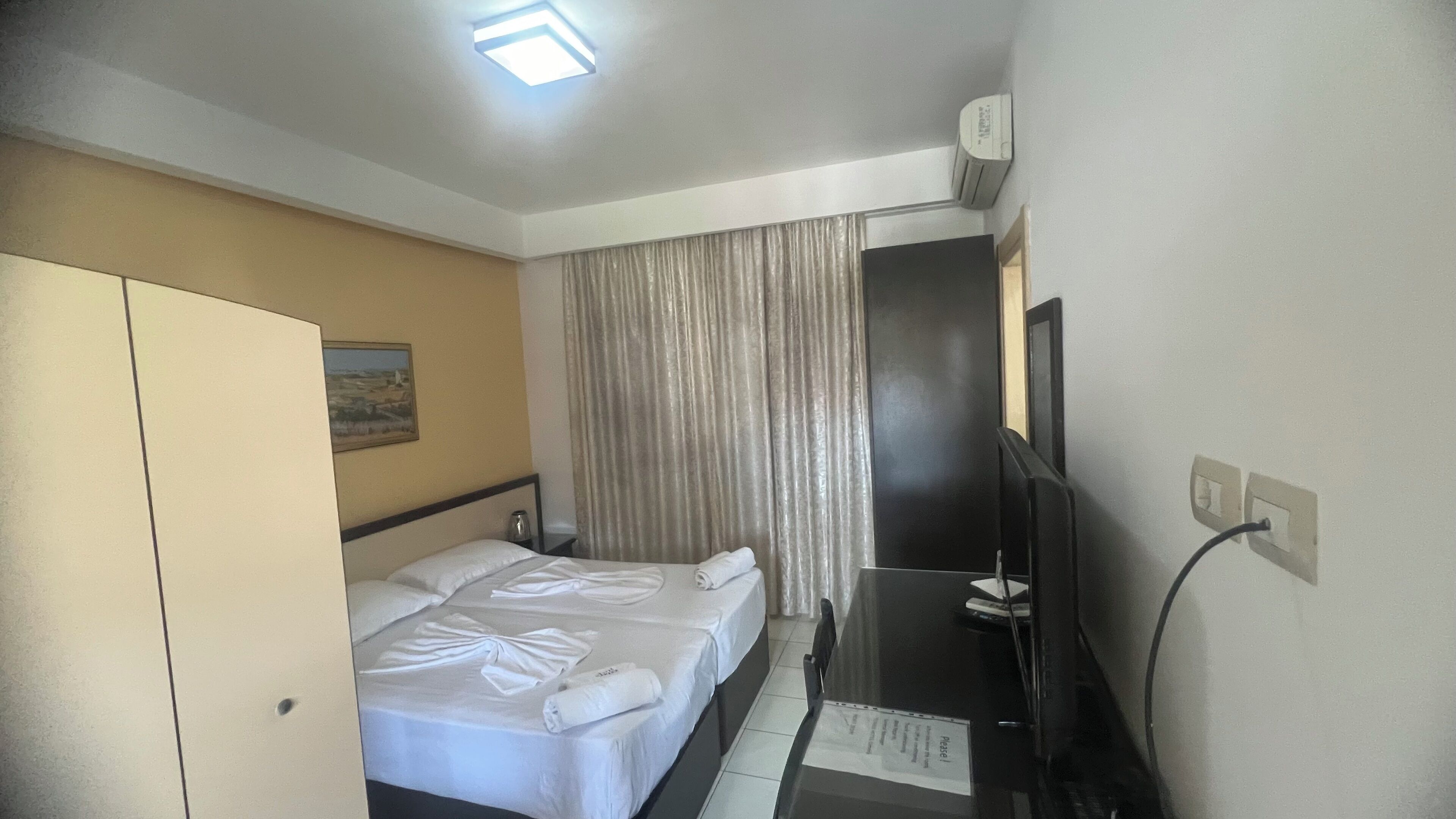 Ein Hotelzimmer mit einem Bett, einem Schreibtisch mit Computer, einem Fernseher und einem Fenster mit Vorhängen.