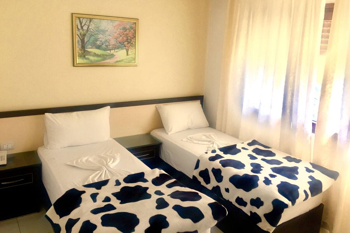 A hotel room with two beds, each with a white bedspread and a cow print blanket. There is a painting on the wall above the beds, and a window with curtains to the right.