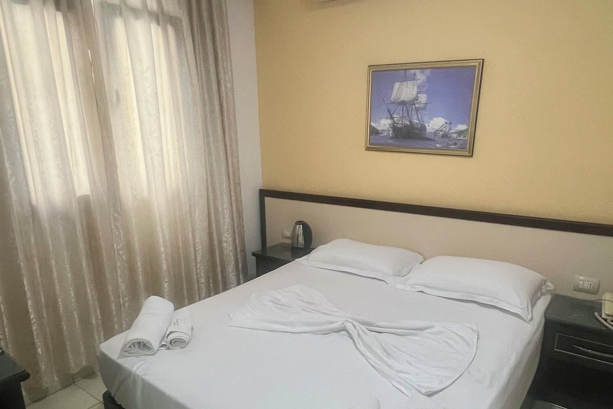 A hotel room with a bed, a nightstand, an air conditioning unit, a painting on the wall, and a window with curtains.