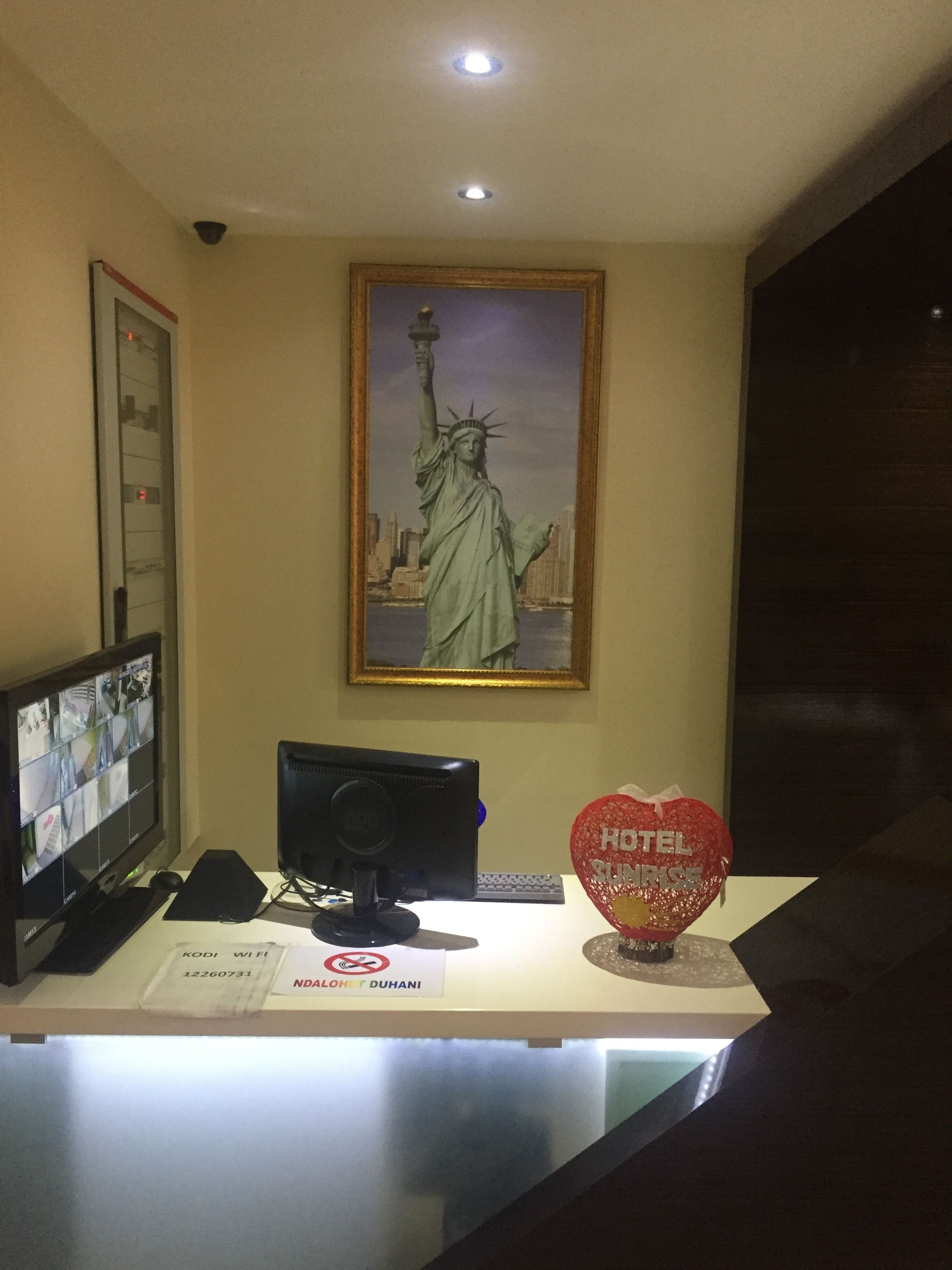Un mostrador de recepción con un monitor que muestra múltiples imágenes de cámaras de seguridad, una estatua enmarcada de la Estatua de la Libertad y un letrero rojo con forma de corazón que dice 'HOTEL'.