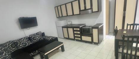 Un espacio habitable compacto con un sofá, una mesita, una kitchenette con armarios y un televisor montado en la pared.