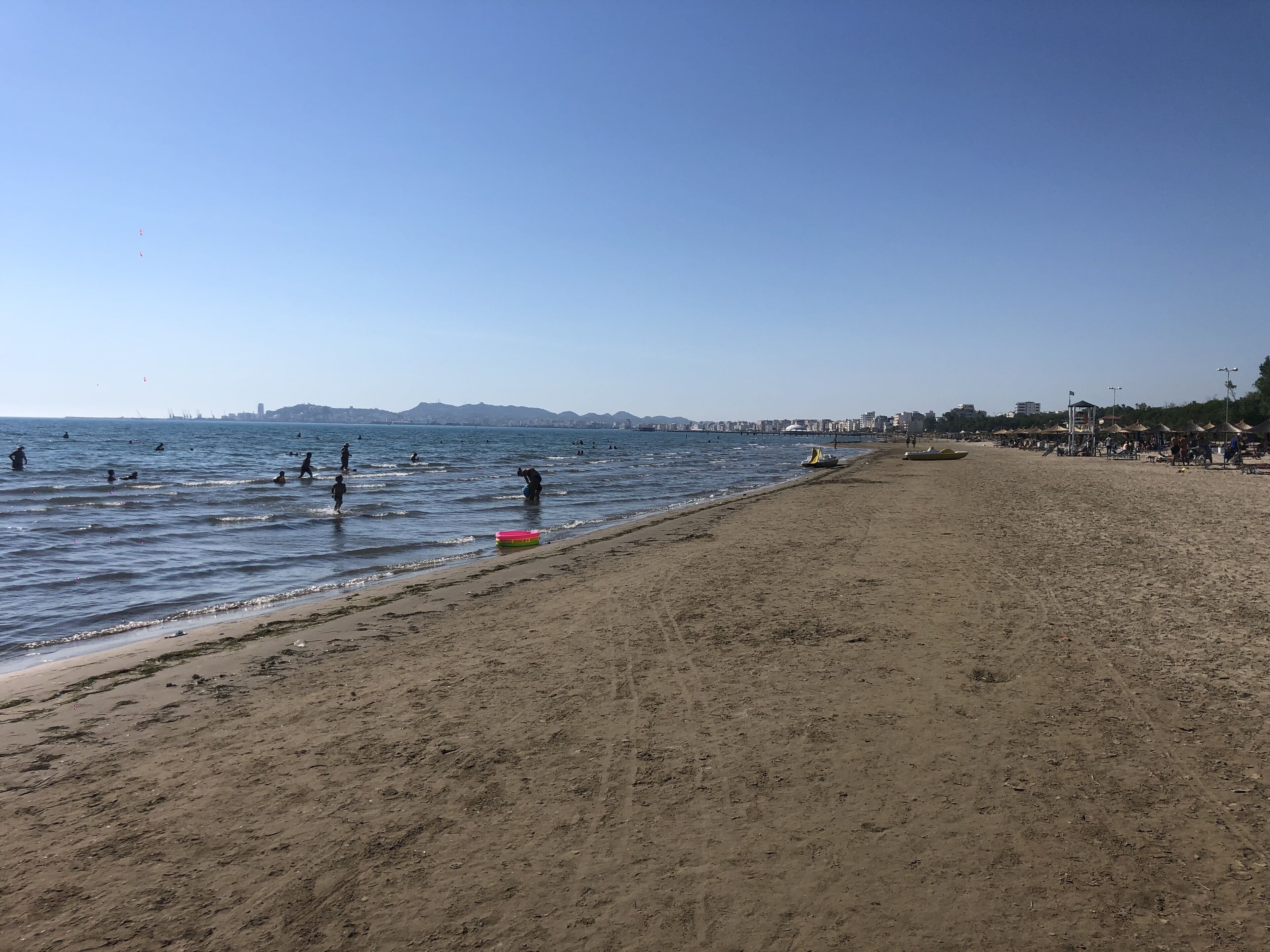A beach with people swimming and playing, a clear sky, and distant buildings.