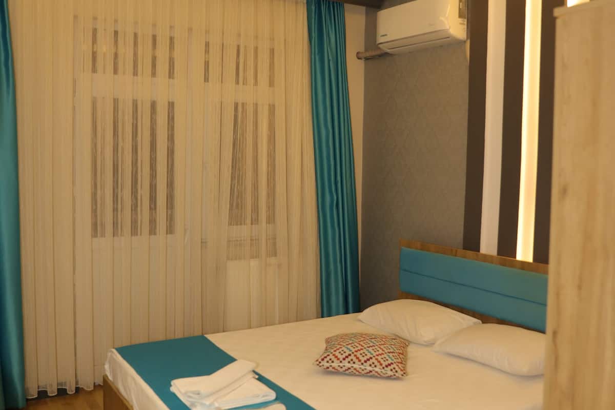 Quarto de hotel com cama de madeira, colcha azul, cortinas brancas e luminária moderna no teto.