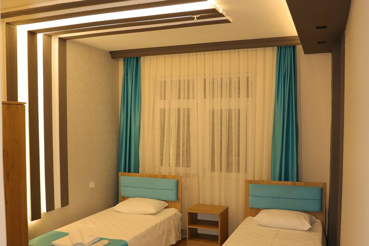 Quarto de hotel moderno com duas camas de solteiro, piso de madeira, mesa de cabeceira e uma janela grande com cortinas.