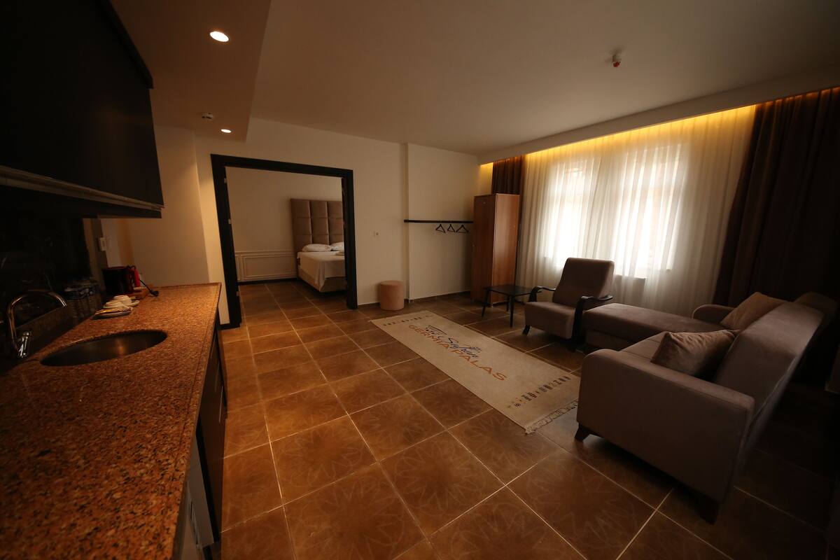 Quarto de hotel moderno com cozinha, sala de estar com sofá e poltronas, e banheiro visível por uma porta aberta.