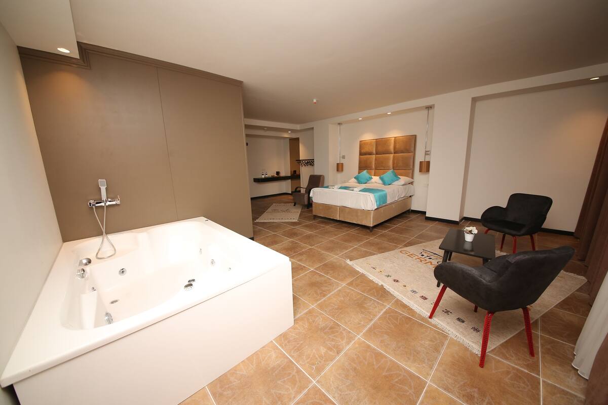 Quarto de hotel moderno com uma banheira branca grande, uma cama com roupa de cama azul, uma mesinha de centro com uma chávena e duas cadeiras pretas.