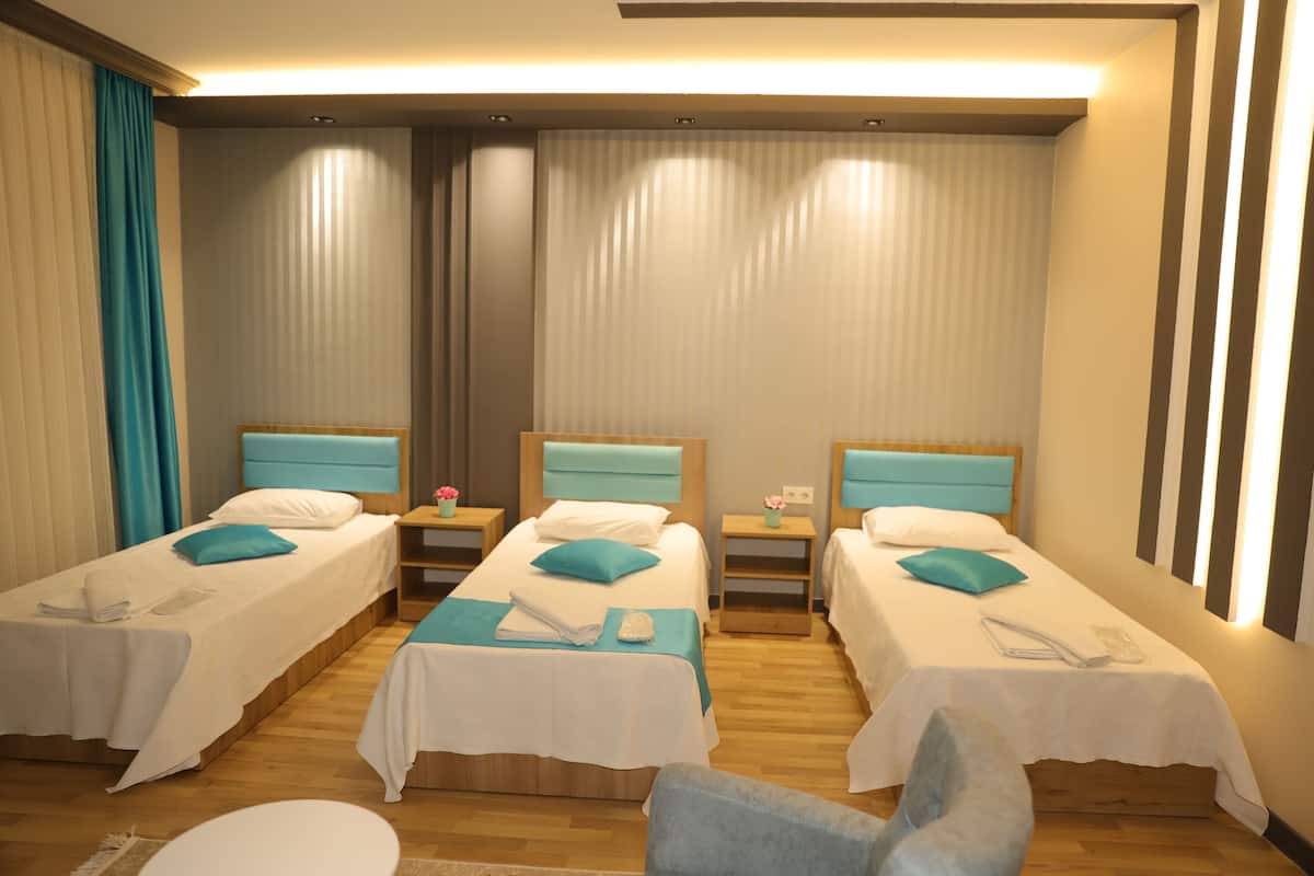 Quarto de hotel com três camas de solteiro, cada uma com colcha branca e almofadas verde-água. Há mesas de cabeceira de madeira com luminárias, uma mesinha e uma cadeira.
