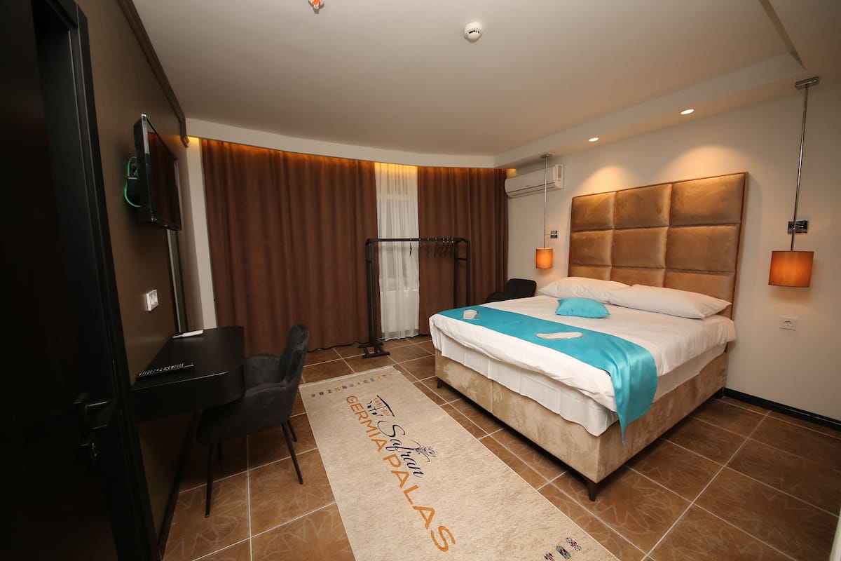 Quarto de hotel com cama, escrivaninha, cadeira e uma janela grande com cortinas.