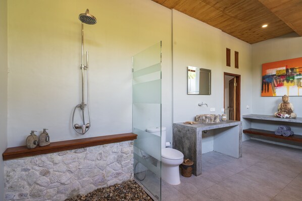 Une salle de bain avec un mur en pierre, une douche en verre, des toilettes blanches et un meuble-lavabo en bois.