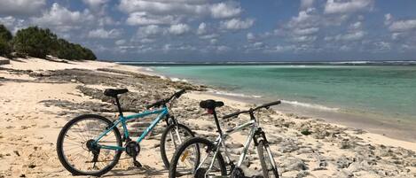 Twee fietsen staan geparkeerd op een zandstrand met helder turquoise water en een bewolkte lucht.