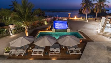 Área da piscina com espreguiçadeiras e guarda-sóis, lareira e vista para o oceano ao entardecer.