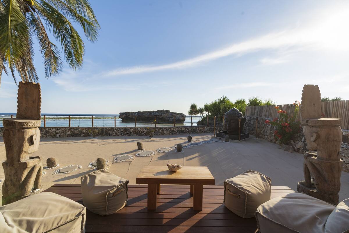 An outdoor patio with a wooden table and beige cushions, overlooking a beach with a rocky outcrop and a wooden fence.