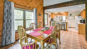 A wooden dining table with red placemats and wooden chairs in a cabin-style room with a kitchen in the background. - New Hampshire Vacation Rental - Walk to Beach! (Madison)