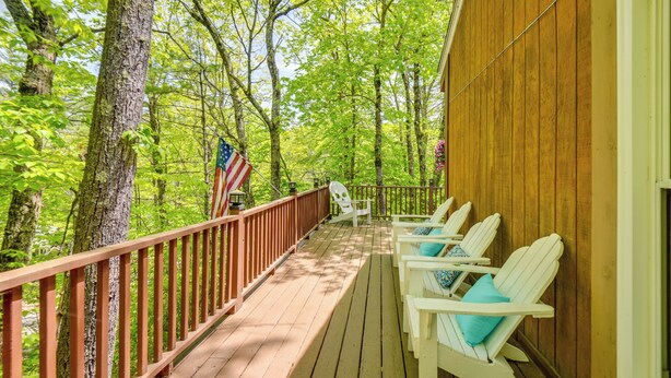 A wooden deck with white chairs and a red railing, surrounded by trees and an American flag. - New Hampshire Vacation Rental - Walk to Beach! (Madison)