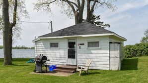 Une petite cabane blanche avec un toit noir, un barbecue, une poubelle bleue et une chaise en bois.