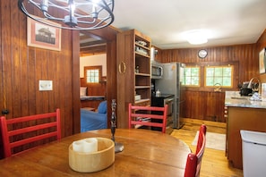 Un intérieur en bois comprenant une table à manger, des chaises, un coin cuisine équipé d’appareils électroménagers et une vue sur l’extérieur.