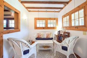 A room with a high ceiling, wooden beams, a window with multiple panes, wicker furniture, and a bookshelf with books.