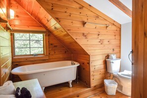 Une salle de bain rustique, avec des murs en bois, comprenant une baignoire sur pieds, des toilettes et une fenêtre.