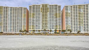 Ein Hochhaus-Hotel mit mehreren Balkonen, ein Strand im Vordergrund und Palmen.