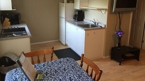 Une cuisine compacte équipée d’un réfrigérateur, d’une cuisinière, d’un évier et de meubles blancs. On y trouve une table à manger avec une nappe à motifs et une chaise. Un téléviseur à écran plat est fixé au mur.