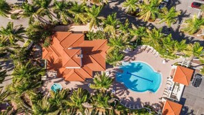Vista aérea de un complejo turístico con piscina, palmeras y un edificio de techo rojo.