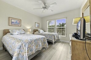 Habitación de hotel con dos camas, ventilador de techo, televisión y ventana con vistas a palmeras.