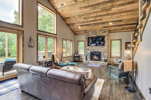 A spacious living room with a stone fireplace, leather sofa, and large windows offering a view of the outdoors. - Modern Lakeside Chalet on Crow Wing Chain w/ Dock! (Nevis)