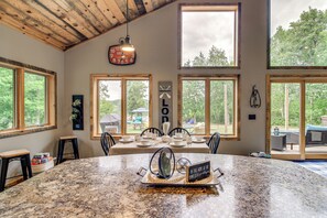 A spacious dining area with a large granite table, multiple chairs, and a view of the outdoors through large windows. - Modern Lakeside Chalet on Crow Wing Chain w/ Dock! (Nevis)