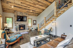 A spacious living room with a stone fireplace, a wooden staircase, and a flat-screen TV mounted above it.