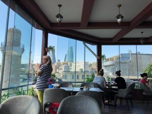 An indoor dining area with large windows offering a city view, tables and chairs, and hanging lights. - Iva Old City Hotel (Baku)