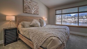 A bedroom with a bed, a nightstand, a lamp, a window with a view of a lake and mountains, and a wall art piece.