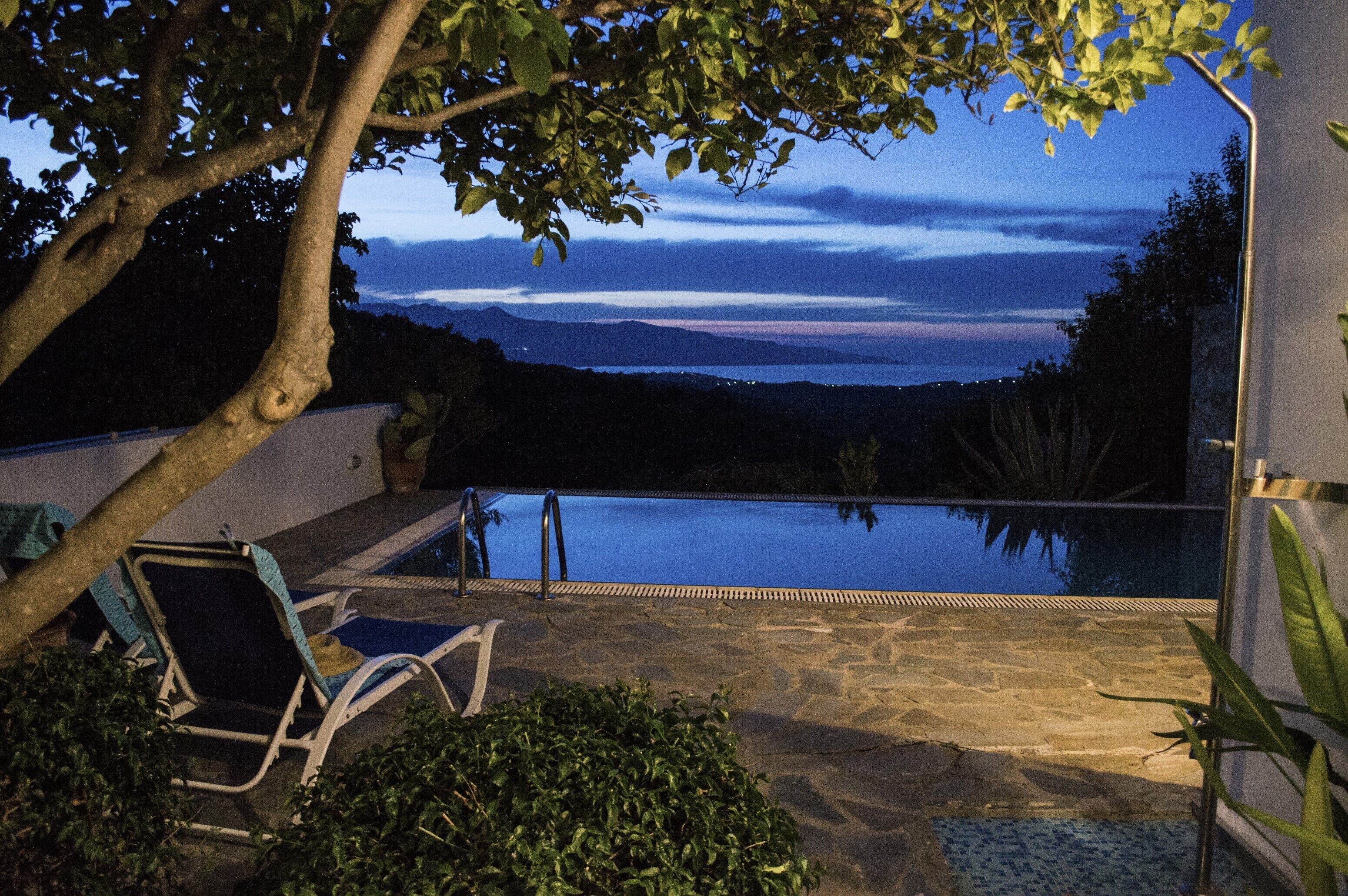 A stone pool with lounge chairs, surrounded by greenery and a view of distant mountains at dusk.