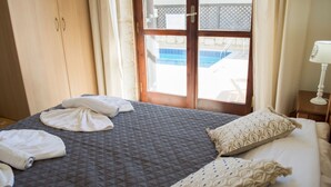 Una habitación de hotel con cama, armario, ventana con vista a una piscina y mesita de noche con lámpara.