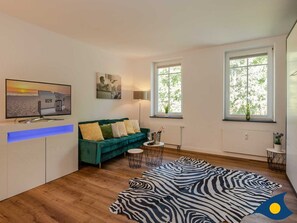 A modern living room with a green sofa, a zebra-patterned rug, a flat-screen TV, and large windows.