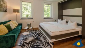 A modern bedroom with a green sofa, a zebra-patterned rug, a bed with white linens, and two windows with a view of greenery. - Holiday Villa Waldstraße Whg. 16 - Holiday Villa Waldstraße Whg. 16 (Heringsdorf)