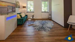 A modern living room with a green sofa, a flat-screen TV, two windows, a zebra-patterned rug, and a small side table with a plant. - Holiday Villa Waldstraße Whg. 16 - Holiday Villa Waldstraße Whg. 16 (Heringsdorf)
