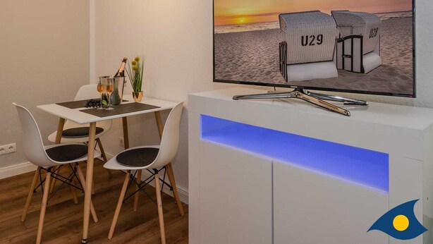 A modern dining area with a TV displaying beach chairs, a table with wine glasses, and a unique ceiling light fixture. - Holiday Villa Waldstraße Whg. 16 - Holiday Villa Waldstraße Whg. 16 (Heringsdorf)
