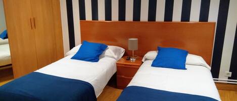Une chambre d’hôtel avec deux lits, chacun agrémenté de draps bleus et d’oreillers blancs. On y trouve une armoire en bois et un mur à rayures derrière les lits.