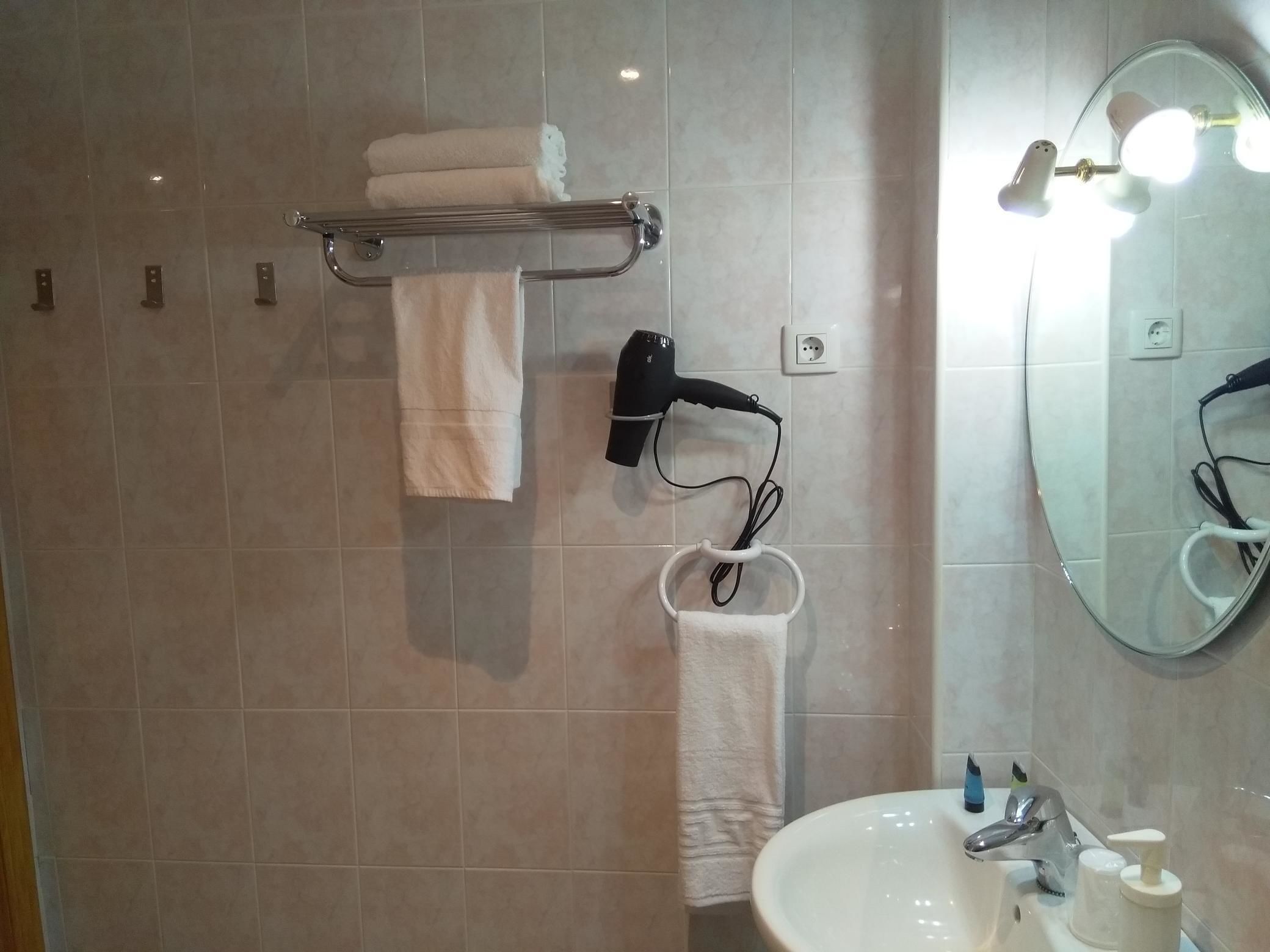 A bathroom with a glass shower enclosure, a towel rack with two towels, a hairdryer, a sink with a mirror, and a soap dispenser.